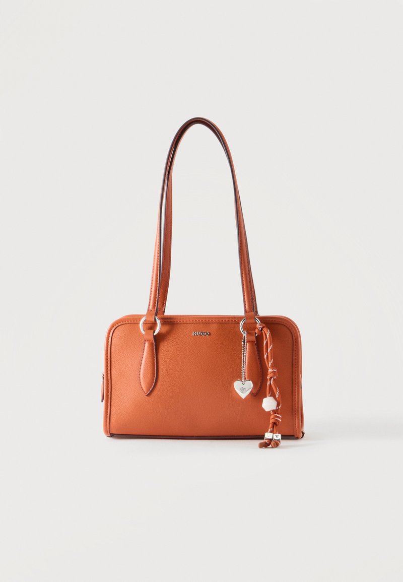 Sac à main en cuir marron avec deux longues anses, un pendentif en métal en forme de cœur et des pompons noués décoratifs sur un fond uni.