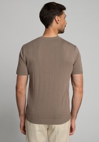 Mężczyzna widziany z tyłu, ubrany w koszulę z krótkim rękawem, prążkowaną, w kolorze taupe oraz jasnobeżowe spodnie na szarym tle.