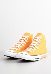 Paire de baskets montantes en toile orange avec semelles en caoutchouc blanc et lacets blancs, posées sur un fond clair et uni.