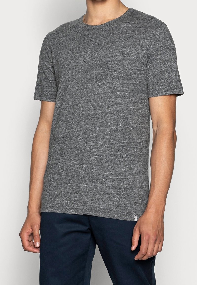 Mand i en kortærmet heather grå crew neck t-shirt og mørkeblå bukser, der står foran en ensartet lys baggrund.