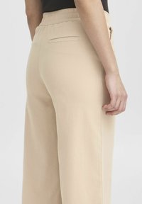 Pantaloni beige con vita elasticizzata e texture liscia, caratterizzati da una singola tasca posteriore e un design a gamba dritta, visibili in una vista laterale.