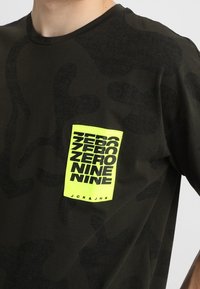 Černé maskáčové tričko vyrobené z jemné tkaniny, s jasně žlutou kapsou a výrazným textem "ZERO NINE" a značkovým štítkem "JCK&JNS".