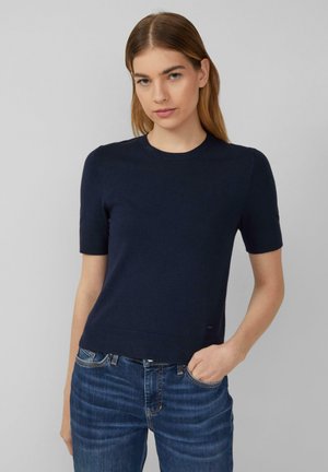 Maglione lavorato a maglia blu navy con maniche corte, scollo rotondo e orlo a costine. Indossato con jeans blu, che mettono in risalto uno stile casual.