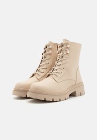 Beige ankelstøvler med en glat læderfinish, rund tå, kraftig sål, snørret front og dekorative metaløjer.