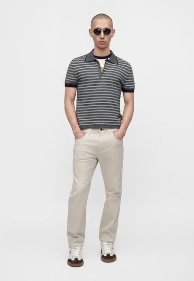Homme à la tête rasée portant des lunettes de soleil noires, un polo rayé gris et blanc, un pantalon beige clair et des baskets blanches avec des détails noirs.