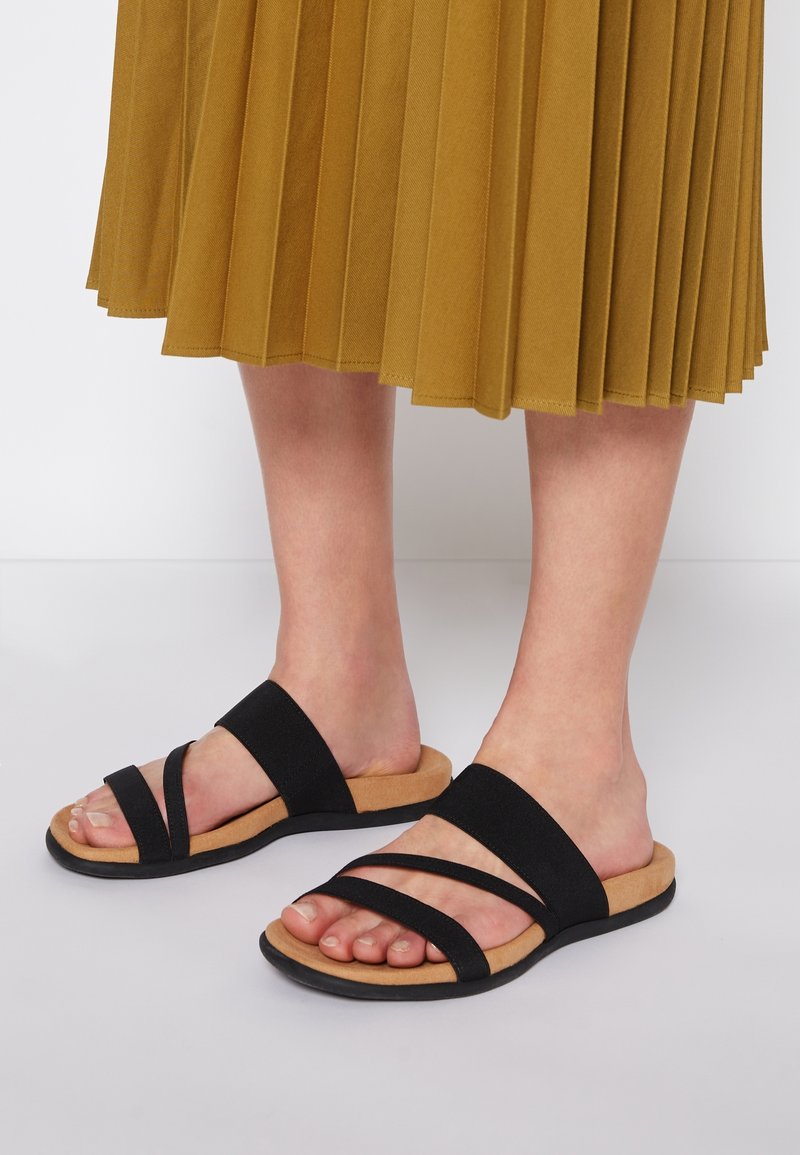 Pieds portant des sandales noires à brides avec des semelles couleur cuir clair, associées à une jupe plissée jaune moutarde sur un fond de sol blanc.