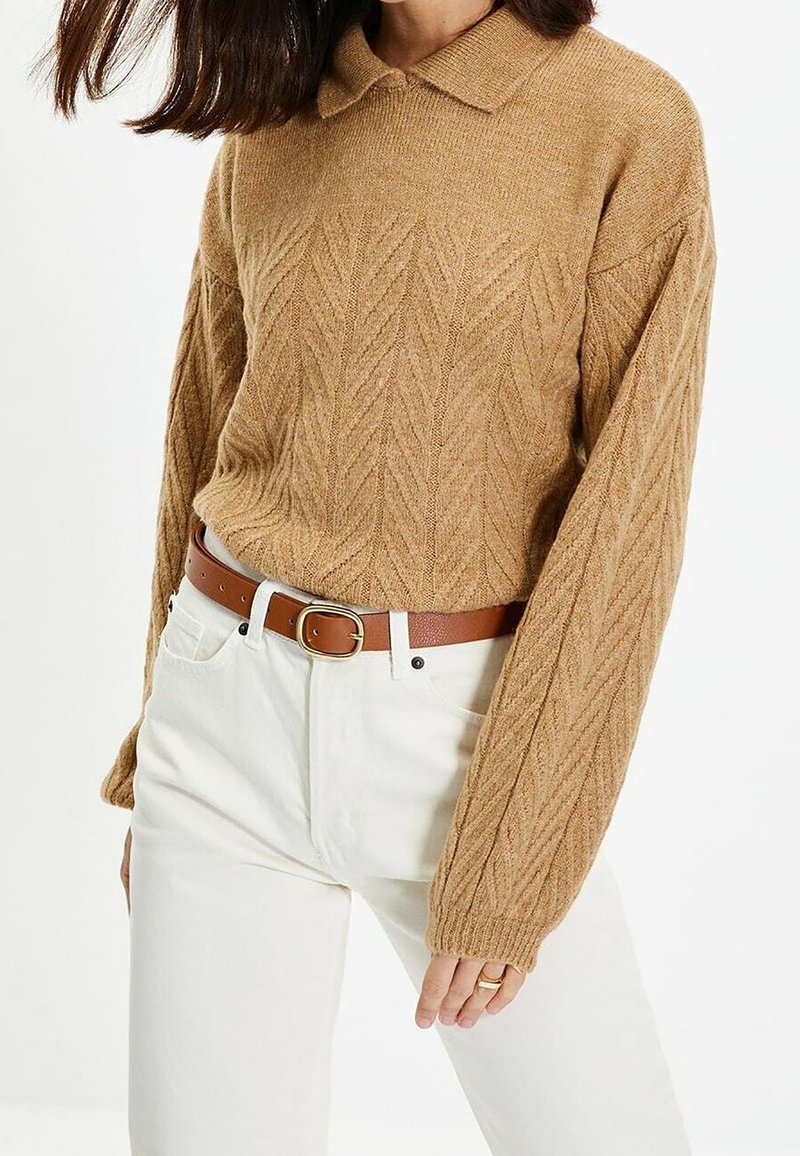 Suéter de punto beige con un patrón de hojas texturizado y un cuello con solapa, combinado con jeans blancos y un cinturón de cuero marrón.
