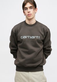 Hanorac maro cu manșete și tiv cu nervuri, având logo-ul "carhartt" de culoare albastru deschis pe parte frontală. Textură netedă, croi lejer, guler rotund.