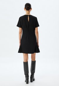 Schwarzes Kleid mit kurzen Ärmeln, ausgestelltem Saum und Rückenschlitz. Kombiniert mit knielangen schwarzen Stiefeln aus glattem Leder.