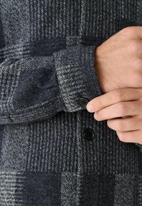 Šedá a černá kostkovaná košile s jemnou texturou, která má černé knoflíky a manžety na knoflíky. Design zahrnuje vodorovné a svislé pruhy.