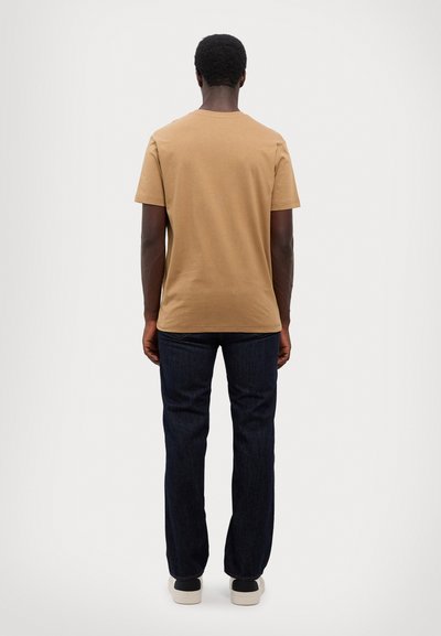 T-shirt en coton marron avec un col rond, manches courtes, associé à un jean en denim bleu foncé, coupe droite, et des baskets blanches et noires.