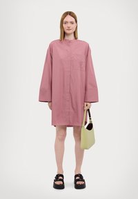 Vestito camicia oversize rosa con colletto, maniche lunghe e taschino sul petto. Il modello tiene in mano una borsa verde chiaro e indossa sandali platform neri.