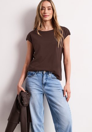 Frau mit langen blonden Haaren, die ein braunes ärmelloses Top und hellblaue Jeans trägt und eine gefaltete dunkelbraune Jacke an ihrer Seite hält.