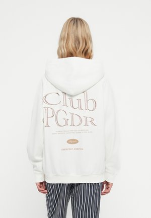 Witte katoenen hoodie met een grote gedrukte afbeelding op de achterkant, met tekst in bruin. Lange mouwen en een voorste buidelzak. Gestreepte broek zichtbaar.