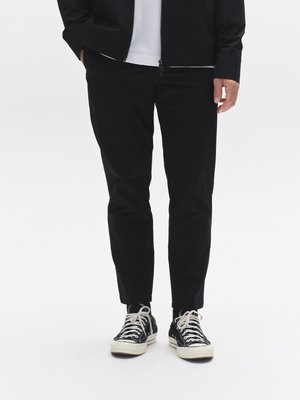 Zwarte taps toelopende broek gemaakt van lichtgewicht stof, gecombineerd met zwarte canvas sneakers. Het jack heeft een ritssluiting en een gladde textuur.