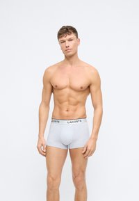 Grå bomulls boxershorts med en svart Lacoste-logo på midjebåndet, med en tettsittende passform og glatt tekstur, vist av en skjorteløs mann.