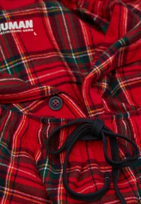 Červená kostkovaná tkanina se zelenými a žlutými akcenty, s knoflíkovým zapínáním a černou šňůrkou. Měkká textura s uvolněným designem.