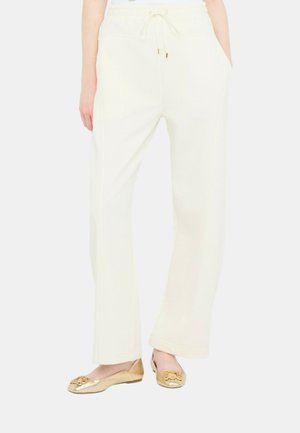 Donna che indossa pantaloni larghi bianco sporco con fascia elastica in vita e scarpe piatte dorate con fibbie decorative.