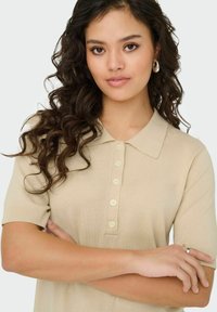 Kreminė trumpomis rankovėmis polo marškinėliai su apykakle, su mygtukų eilučių ir raukšlėtu tekstūra. Modelis turi garbanotus plaukus ir peržengtas rankas.