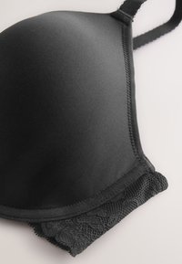 Bonnet de soutien-gorge rembourré noir avec tissu lisse et bordure en dentelle sur le bord inférieur sur un fond neutre.