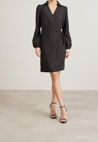 Robe noire cintrée à manches bouffantes longues, décolleté en V profond, taille cintrée, lien décoratif et ourlet asymétrique, portée avec des talons noirs à lanières.
