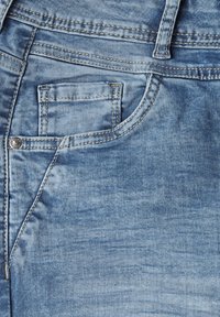 Jean en denim bleu clair avec une poche avant, des coutures visibles et une texture usée présentant des variations subtiles de couleur.