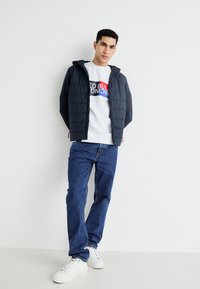 Granatowa kurtka puchowa z kapturem, noszona na białym swetrze z wielokolorowym logo, w zestawieniu z niebieskimi dżinsami i białymi trampkami.