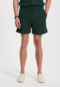 Pantaloni scurți de culoare verde închis, texturați, cu model în tapițerie și talie elastică cu şnur. Asortați cu pantofi slip-on de culoare crem.