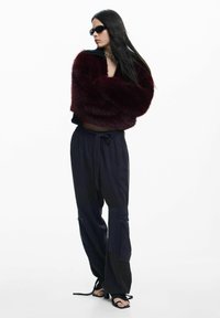 Bordová krátka bunda z umelého kožušina, čierne nohavice sťahované šnúrkou a so zipsovými vreckami, čierne sandále na remienky a tmavé slnečné okuliare.