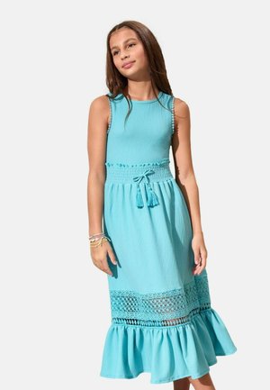 Dievča stojace, oblečené v svetlomodré midi šaty bez rukávov so stiahnutým pásom, strapcovým šnúrkou, čipkovaným lemom a volánovým spodkom, na bielom pozadí.