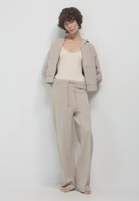 Donna con capelli ricci che indossa pantaloni a gamba larga beige, una felpa cropped con zip abbinata e una canotta bianco sporco, scalza contro un muro semplice.