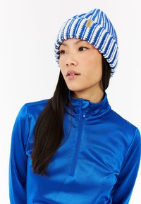 Pletená čepice se modro-bílými pruhy, s texturovaným designem a malou štítkovou značkou. Model má na sobě modrou zipovou bundu.