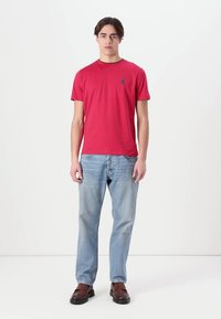 Rød kortermet t-skjorte med rund hals og en liten logo på brystet, kombinert med lyseblå jeans og brune sko.