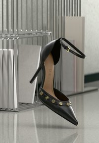 Schwarzer hoher Schuh mit spitzem Zehenbereich, Knöchelriemen und goldenen Zierakzenten entlang des Vorderteils. Hergestellt aus glattem Leder. Markenlogo sichtbar.