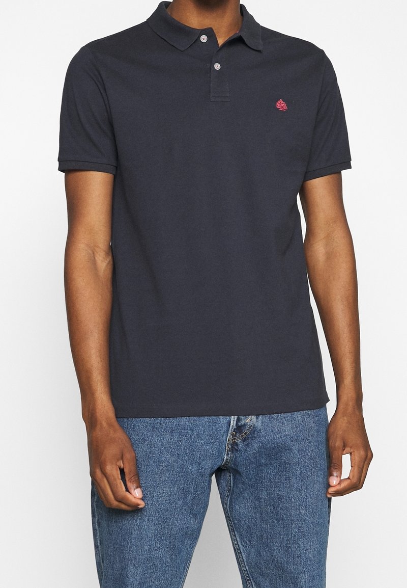 Polo shirt navy en coton avec un col classique et des manches courtes, arborant un petit logo rouge sur la poitrine gauche. Porté avec un jean en denim bleu.