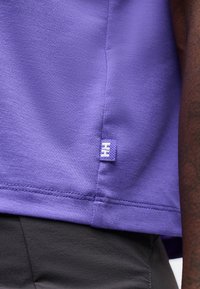 Κοντινό πλάνο στο τελείωμα μοβ υφάσματος με μια μικρή υφασμένη ετικέτα, φορεμένο πάνω από σκούρο παντελόνι, που δείχνει την υφή του υφάσματος και τις λεπτομέρειες ραφής.