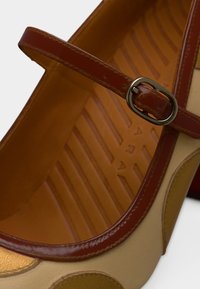 Chaussure en cuir beige avec un bout rond et une bride marron dotée d'une boucle en métal. L'intérieur révèle une semelle texturée et striée.