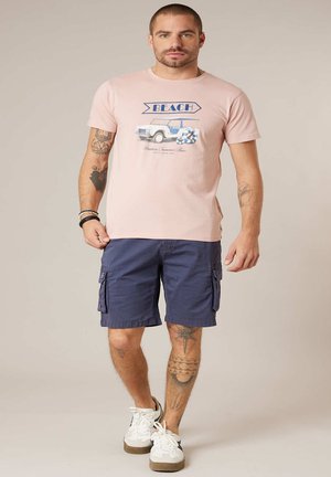 Homme avec des tatouages portant un t-shirt rose à motif "BEACH", un short cargo bleu marine, des baskets blanches et des bracelets, marchant devant un fond uni.
