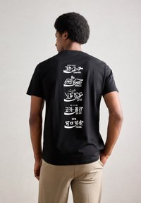 Black t-shirt featuring multiple white logos in various scripts on the back. Short sleeves and a relaxed fit. Neutral background.