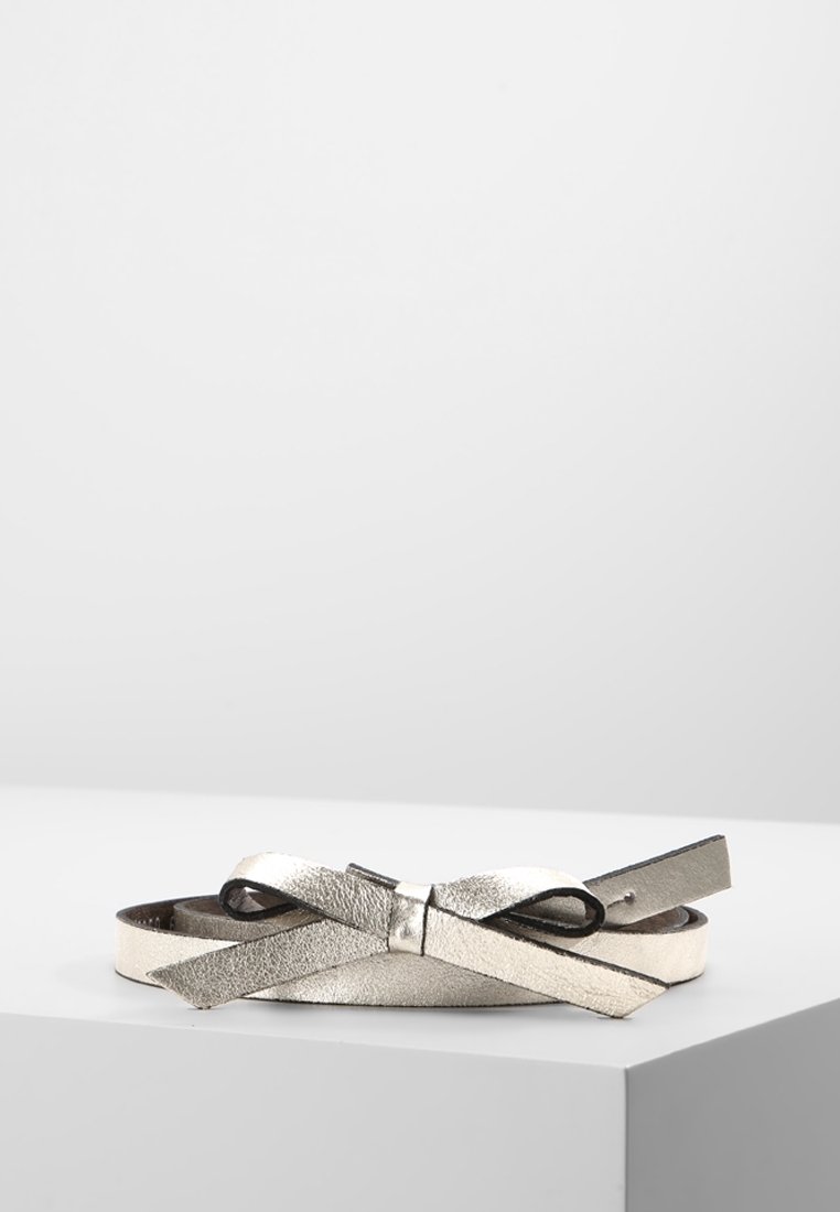 Ceinture fine en cuir argenté métallique avec un petit détail en forme de nœud, enroulée et posée sur une surface blanche sur fond gris uni.