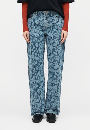 Osoba koja nosi plave široke hlače visokog struka sa svijetloplavim cvjetnim uzorkom, crne cipele i crnu majicu s crvenim dugim rukavima.