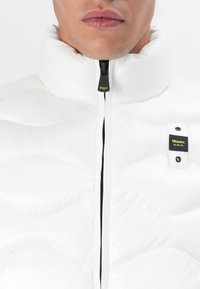 White puffer jacket with high collar, black zipper, and a visible logo patch on the chest. Features quilted texture and sleek design.