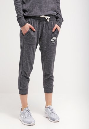 Personne portant un pantalon de survêtement Nike gris foncé avec des poches et un cordon blanc, associé à des baskets Nike gris clair, debout sur un fond blanc.
