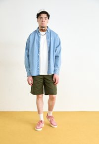 Lichtblauw denim overhemd met knopen over een wit T-shirt, gecombineerd met olijfgroene shorts en roze sneakers. Geplaatst tegen een effen achtergrond.