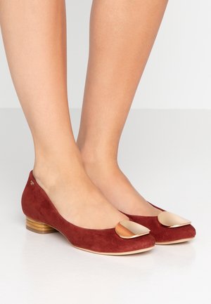 Røde ruskinds-balletsko med en guldfarvet detalje på tåen, præsenteret på en glat hvid overflade, der fremhæver skoens elegante design.