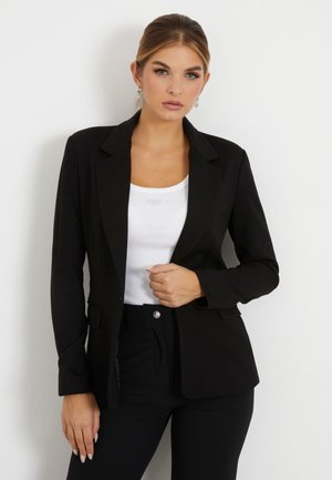 Blazer nero aderente con rever a punta, due tasche frontali e una texture liscia. Indossato sopra un top bianco, abbinato a pantaloni neri.