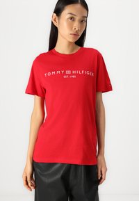 Red cotton T-shirt featuring a white logo "TOMMY HILFIGER EST. 1985" on the front. Short sleeves, round neck, and relaxed fit.