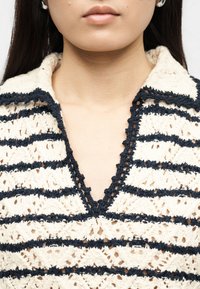 Γυναίκα που φοράει επάνω μέρος από κρεμ και ναυτικό μπλε πλεκτό βελονάκι με γιακά σε σχήμα V, αποκαλύπτοντας τον λαιμό και το κάτω μέρος του προσώπου της.