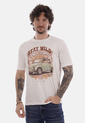 Uomo con capelli ricci e tatuaggi che indossa una maglietta bianca con grafica di un'auto d'epoca, raggi di sole, fiori e testo "Stay Wild".