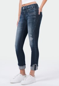 Donkerblauwe denim jeans met een slim fit, verwerkte details, franje aan de onderkant en zilveren hardware accenten. Gestyled met witte sneakers.